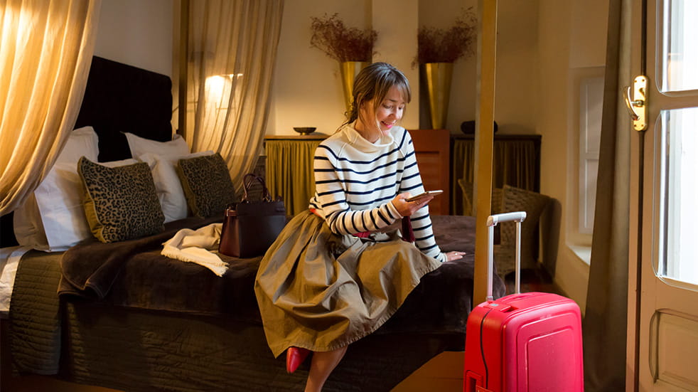 Woman sitting on hotel bed, text messaging on mobile phone